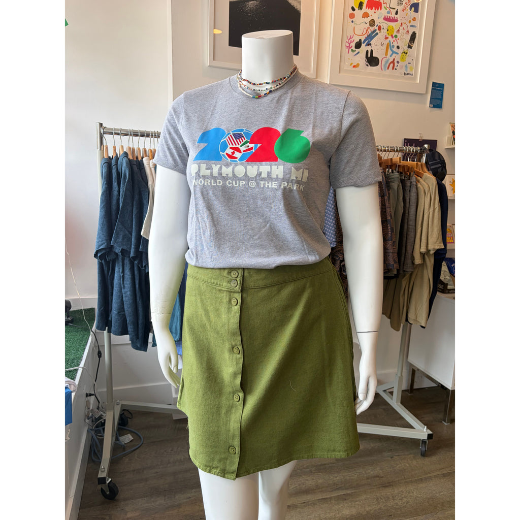 A gray graphic t-shirt with a colorful '2025 World Cup' design paired with an olive green button-up skirt, displayed on a white mannequin inside a clothing store.